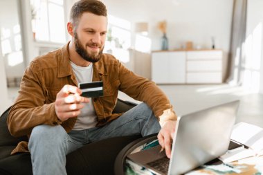 Genç bir adam bir elinde kredi kartı tutuyor ve bilgisayarını parlak, havadar bir oturma odasında kullanıyor. Çevrimiçi alışverişe odaklanmış ve meşgul görünüyor..