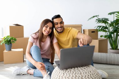 Mutlu bir çift, açık kutularla çevrili, parlak bir oturma odasında yumuşak bir halıya oturur. Bir dizüstü bilgisayara odaklanırlar, gülümsemelerini paylaşırlar ve birlikte anın tadını çıkarırlar..