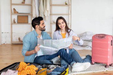 Genç bir çift, seyahat çantaları ve giysileriyle çevrili bir şekilde yerde oturmuş, gülümsüyor ve haritaya bakıyor. Yaklaşan seyahatlerini planlamak için heyecanlı görünüyorlar..