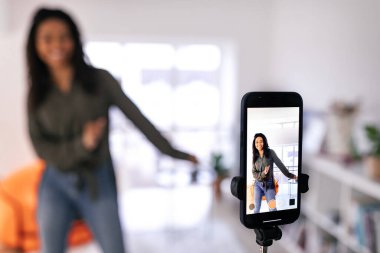 Neşeli genç bir kadın, rahat bir oturma odasında telefonuyla video kaydederken dans ediyor. Parlak alanda rahat mobilyalar ve pencereden gelen doğal ışık yer alıyor..