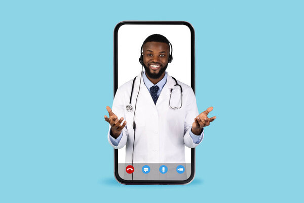 Онлайн медицинская консультация. Симпатичный черный доктор в наушниках, выглядывающий из большого экрана смартфона, изолированный на синем фоне, афроамериканский врач в униформе, жест перед камерой, коллаж