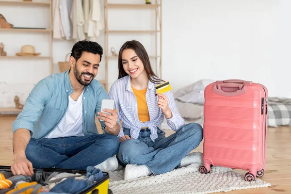 Bir çift, etrafları bavullar ve seyahat eşyalarıyla çevriliyken seyahat planlarını heyecanlı bir şekilde tartışıyor. Konforlu, aydınlık bir yerde rezervasyon yaptırmak için telefon ve kredi kartı kullanıyorlar..