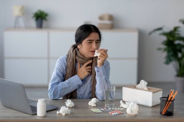 Genç Hintli bir kadın ofis masasında kendini kötü hissederek oturuyor. Bir peçeteye öksürüyor ve etrafı peçetelerle, haplarla ve bir bardak suyla çevrili. Bu da iş yerindeki hastalığın mücadelesini gösteriyor..