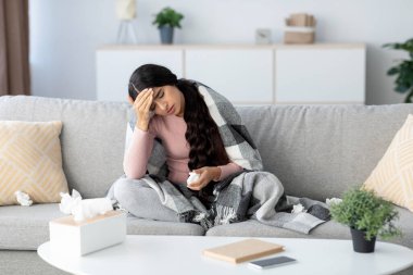 Genç Hintli bayan battaniyeye sarılı olarak kanepede oturuyor. Grip ve baş ağrısı belirtileri gösteriyor. Etrafa saçılmış dokularla ilaç tutuyor. Hastalıkla mücadele ettiğini gösteriyor..