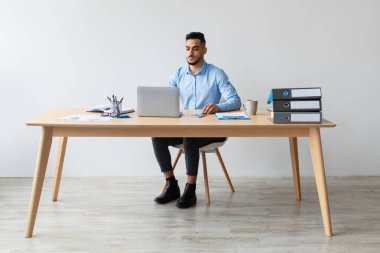 Orta Doğulu erkek yönetici, modern bir masada dizüstü bilgisayarı üzerinde çalışıyor. E-postaları kontrol ediyor ve gündüz vakti parlak bir ofiste web sitesine katılıyor..