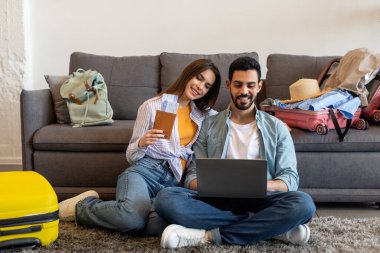 İki kişi yerde oturuyor, etrafı bavullarla çevrili. Kadın uçuş biletlerini tutarken ve ortağına gülümserken mutlu bir şekilde seyahatlerini planlıyorlar..