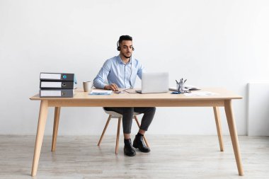 Orta Doğulu bir adam modern bir ofiste masa başında oturuyor. Kulaklık takıyor ve müşterilerine danışmak için bilgisayar kullanıyor. Profesyonel bir iş ortamı yaratıyor..