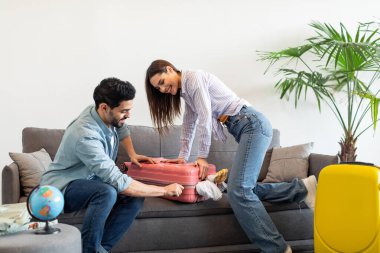 Bir çift, seyahat için hazırlanırken pembe bir bavulu kapatmaya çalışıyor. Oturma odası parlak ve kanepenin yanında yeşil bir bitki var. Heyecanları çok açık..