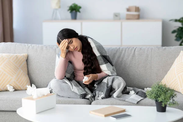 Genç Hintli bayan battaniyeye sarılı olarak kanepede oturuyor. Grip ve baş ağrısı belirtileri gösteriyor. Etrafa saçılmış dokularla ilaç tutuyor. Hastalıkla mücadele ettiğini gösteriyor..