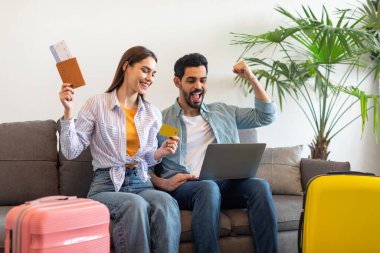Mutlu çift koltukta oturup tatil rezervasyonlarını kutluyor. Samimi bir odada bitkilerle dolu bir dizüstü bilgisayara bakarken ellerinde biletler ve bir kredi kartı var..