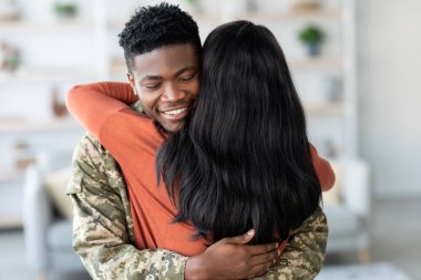 Askeri üniformalı bir asker samimi bir oturma odasında partneriyle samimi bir kucaklaşmayı paylaşır. Sahne aşkı, mutluluğu ve sıcak bir atmosferde neşeli bir kavuşmayı yansıtıyor..