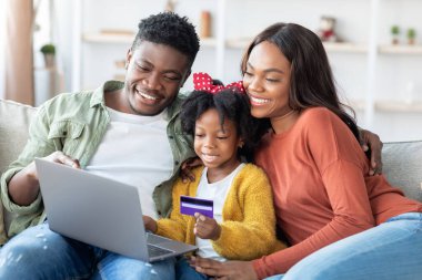 Neşeli bir aile, parlak bir oturma odasında bir kanepede oturur. Online alışveriş yapıyorlar, ellerinde bir kredi kartı olan ve gülümseyen çocukla. Hava sıcak ve davetkar..