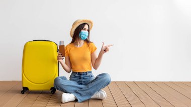 Maske takan ve şapka takan genç bir kadın, sarı bir bavulun yanında, ahşap bir zeminde oturuyor. Bir içki tutuyor ve işaret ediyor, muhtemelen yaklaşan bir seyahat noktasını işaret ediyor..