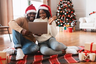 Noel Baba şapkalı neşeli bir siyah çift evde yerde oturup Noel için bir video görüşmesi yapıyor. Tatil süslemeleri ve hediyelerle çevrili iken heyecanla ellerini sallıyorlar..