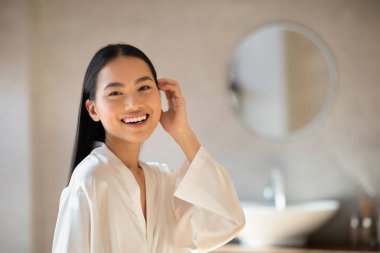 Modern bir banyoda, beyaz bornozlu genç bir Japon bayan uzun ipeksi saçlarına dokunurken kameraya gülümsüyor..