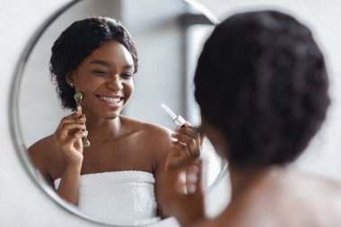 Parlak bir banyoda, genç bir Afrikalı Amerikalı kadın aynanın önünde duruyor. Sabah duşundan sonra cilt bakımı ve neşe saçan rutini için yüz silindiri ve serum kullanıyor..