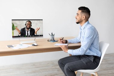 Gülümseyen genç bir Arap, ahşap bir masada dizüstü bilgisayar ve defterle oturuyor. Parlak bir ofisin uzaktan kumandası sırasında bir meslektaşıyla görüntülü konuşma yapıyor..
