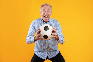 Heyecanlı, olgun bir adam elinde futbol topu tutan, parlak sarı bir arka plana karşı duruyor. En sevdiği takımı desteklerken eğlenceli bir oyuna hazır olduğunu gösteriyor..