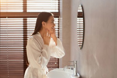 Uzun saçlı güzel bir kadın aynadaki yansımasına bakarken gülümsüyor. İpek bornoz giyiyor ve penceresi olan iyi aydınlatılmış bir banyoda..