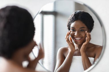 Havluyla gezen mutlu, genç bir zenci kadın aynanın önüne göz tedavisi ürünü sürerken gülümsüyor. Evde serinletici bir duştan sonra sabah güzellik rutininin tadını çıkarıyor..