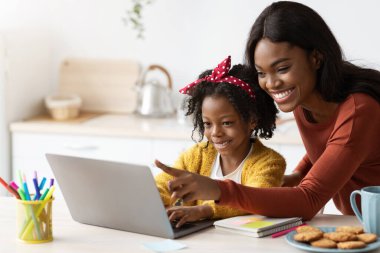 Bir anne ve küçük kızı bir dizüstü bilgisayarla iletişim kurarken neşeli bir anı paylaşıyorlar. Renkli kırtasiye ve atıştırmalıklarla çevrili, parlak bir mutfak masasında oturuyorlar..