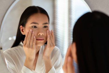 İyi aydınlatılmış bir banyoda, beyaz bornoz giyen genç bir kadın aynada tenini muayene ediyor. Yaşlanmanın ilk belirtilerini vurgulayarak cilt sorunlarıyla ilgili üzüntüsünü ifade ederken yanaklarına dokunuyor..