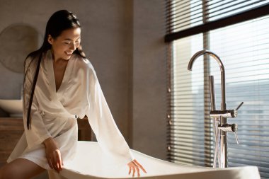 Modern bir küvetin kenarında oturan ve suyun sıcaklığını ölçerken gülümseyen binlerce yıllık mutlu Japon bayan..