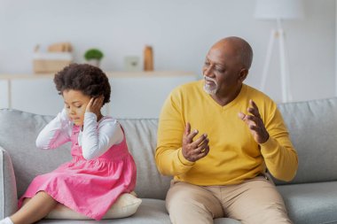 Oturma odasında, küçük bir kız elleri kulaklarının üzerinde oturur, yaşlı bir adam yakınlarda hareketli bir şekilde konuşurken üzüntü ve hayal kırıklığını gösterir. Şu anda aile gerginliği çok açık..