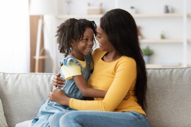 Bir anne ve kızı kanepede birlikte oturur, samimi bir oturma odasında gülüşmeler ve kahkahalar atarlar. Güneş ışığı boşluğu doldurur ve sıcak bir atmosfer yaratır..