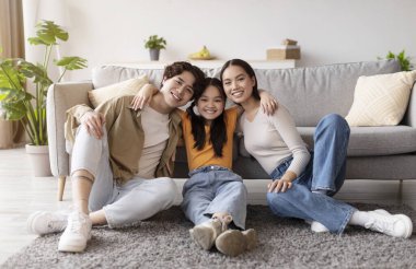 Üç aile üyesi parlak bir oturma odasında yumuşak bir halının üzerinde birlikte oturuyorlar. Çiçekler ve rahat bir kanepe ile çevrili, gülücükler ve kahkahalar paylaştıkça atmosfer sıcak ve neşeli..