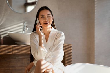 İpek bornozlu neşeli bir bayan duştan sonra telefonla konuşurken gülümsüyor. Güneş ışığı banyoya akar ve sabah rutini sırasında sıcak, davetkar bir atmosfer yaratır..