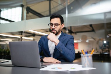 Mavi takım elbiseli bir adam, çağdaş bir ofiste dizüstü bilgisayarına odaklanır. Derin düşüncelere dalmış, muhtemelen verileri analiz ediyor ya da belgeleri inceliyor. Parlak doğal ışık boşluğu doldurur..