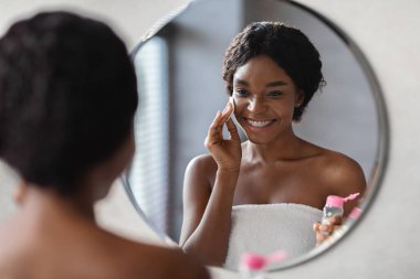 Beyaz havluya sarılı neşeli siyah bir bayan makyaj malzemelerini çıkarmak için pamuk pedin üzerinde mikiler suyu kullanıyor. Aynada kendine bakıyor, rahatlatıcı bir duşa hazırlanıyor..