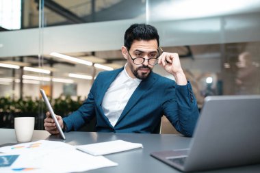 Bir işadamı tabletindeki belgeleri okumaya odaklanmış durumda. Gözlük takıyor, modern bir ofis masasında oturuyor ve yakınlarda dizüstü bilgisayarı ve kahve fincanı var. Doğal ışık odayı aydınlatır..