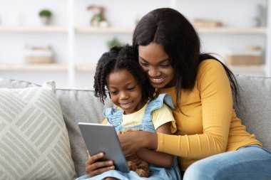 Neşeli bir anne ve kızı bir kanepede oturup tabletle etkileşime girerken neşeli bir anı paylaşıyorlar. Doğal ışıkla dolu parlak bir oturma odasındalar..