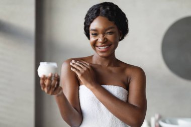 Modern bir banyoda parlak bir sabah, gülümseyen genç siyah bir kadın koluna vücut kremi sürüyor. Beyaz bir havluyla kaplanarak, ferahlatıcı bir duştan sonra cilt bakımını sever..