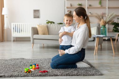 Bir anne, parlak bir oturma odasında yanında duran genç oğluna diz çöküp gülümsüyor. Renkli oyuncaklar aralarındaki yumuşak halıya saçılmış neşeli bir atmosfer yaratıyorlar..