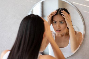 Uzun saçlı Çinli bir kadın banyo aynasında kendine bakıyor ve saçlarının incelmesinden endişe ediyor. Omzunun üstünden bakarken saçlarını gri iplikler için kontrol eder..