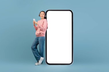 Neşeli genç Asyalı bir kadın boş ekranlı dev bir cep telefonunun yanında duruyor. Kulaklık takıyor ve akıllı telefonunu eğlenceli bir mobil uygulamayı keşfetmek için kullanıyor..
