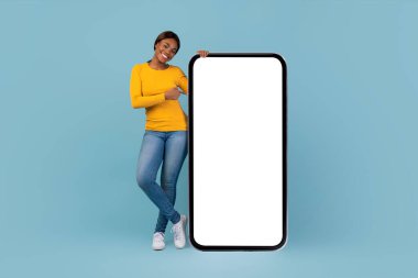 Açık mavi bir arkaplanda duran bir kadın büyük boş bir akıllı telefon ekranını işaret ederken gülümsüyor. Sarı uzun kollu bir gömlek ve kot pantolon giyiyor. Neşeli bir atmosfer yaratıyor..