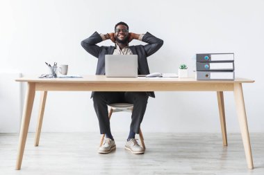 Genç siyah iş adamı ellerini başının arkasına koyarak rahat bir şekilde ofisinde oturuyor. Bilgisayarını temiz beyaz bir arka plana sahip modern bir çalışma alanında kullanırken mola vermeyi seviyor..