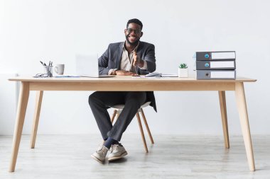 Takım elbiseli genç bir siyahi işadamı, elinde dizüstü bilgisayar ve belgelerle ofisinin masasında güvenle oturuyor..