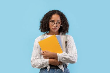 Üzgün siyah bir kız öğrenci elinde birkaç defter tutarken yorgun hissederek mavi bir arkaplanda duruyor. Üniversite sınavlarına ve ödevlerine hazırlanırken yorgunluk belirtileri gösteriyor..