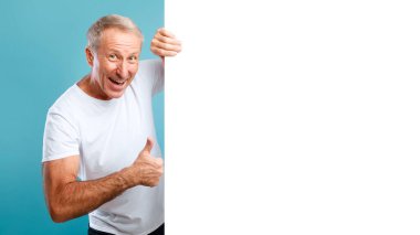 Neşeli, olgun bir adam mavi bir duvara yaslanmış, boş beyaz bir reklam panosundan dışarı bakıyor. Geniş bir gülümseme sergiliyor ve tasarım için dikkat çekiyor..