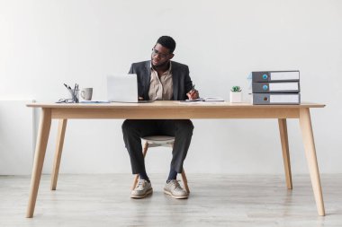 Ciddi bir Afro-Amerikan işadamı masasında dizüstü bilgisayar kullanırken defterine yazmaya odaklanır. Basit bir stüdyoda çevrimiçi eğitim oturumuna katılıyor..