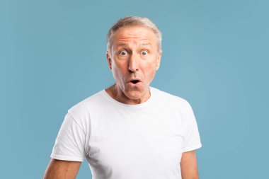 Olgun bir adam mavi arka planlı bir stüdyoda duruyor, şaşkın ve şaşkın bakıyor. Ağzı güvensizlik içinde, beklenmedik haberlere duygusal bir tepki veriyor..