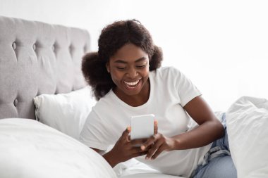 Aydınlık bir sabahta pijamalı neşeli siyah bir kadın cep telefonunu sohbet etmek için kullanırken yatağına oturur ve gülümser. Sıcak yatak odası sıcak ve rahat bir atmosfer yaratıyor..