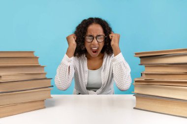 Siyahi bir kız öğrenci üniversite sınavlarına çalışırken hayal kırıklığını ve yorgunluğunu ifade eden kitaplarla dolu bir masada oturuyor. Mavi arka plan, onun stres ve kararlılığını vurguluyor..