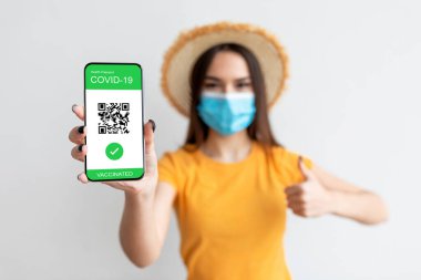 Sarı gömlekli ve hasır şapkalı genç bir kadın elinde akıllı telefonuyla COVID-19 aşı pasaportunu gösteriyor. Onaylıyor, onayını veriyor..