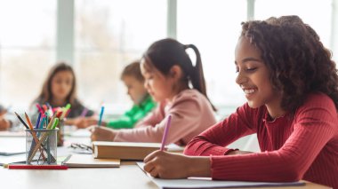 Siyahi bir liseli kız masasında oturuyor ve çok kültürlü sınıf arkadaşlarıyla birlikte parlak, modern bir sınıfta mutlu bir şekilde yazı yazıyor. Öğrenme faaliyetlerine katılıyorlar, görevlerine odaklanıyorlar..